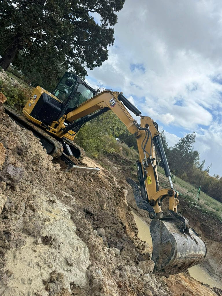 Yellow excavator on a hillside with boom arm extended, digging into earth and dirt mound with cloudy sky background