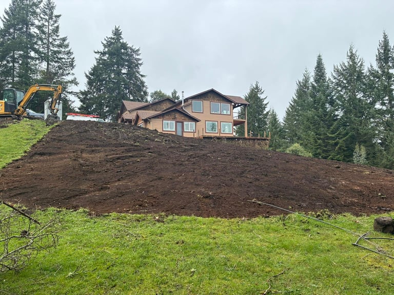 Residential home on hillside with large earthen mound in foreground, yellow excavator, and evergreen forest backdrop