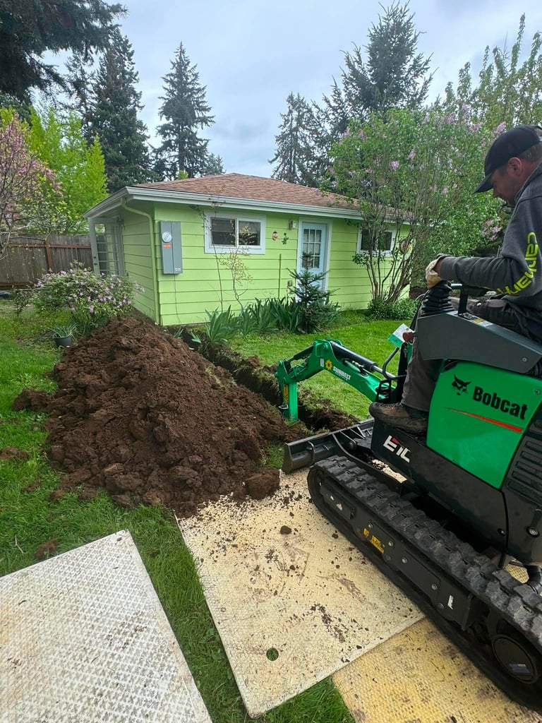Man operating green Bobcat skid steer loader dumping soil in residential yard with small lime green shed in background