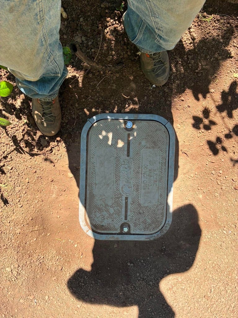 Overhead view of a person standing on bare soil next to a metal utility box cover, casting shadows in sunlight